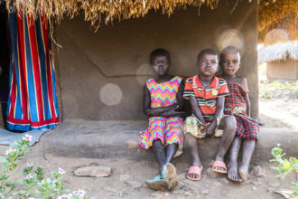 Kolme lasta värikkäissä vaatteissa istuu savimajan edessä Bidibidin pakolaisalueella Ugandassa.