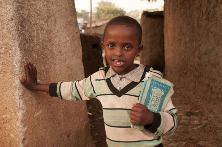 Etiopialainen pieni poika raidallisessa paidassa nojaa seinään kirjoja kainalossaan.