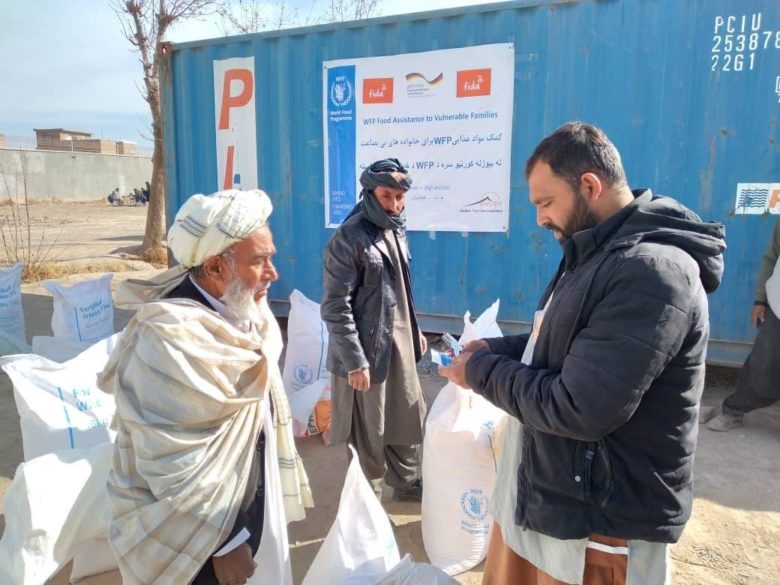 Kuvassa kolme miestä Afganistanissa. Yksi jakaa toiselle ruokakassia avustuksena. Vanha mies ottaa kassia vastaan. Taustalla ohikulkija katsoo tilannetta.