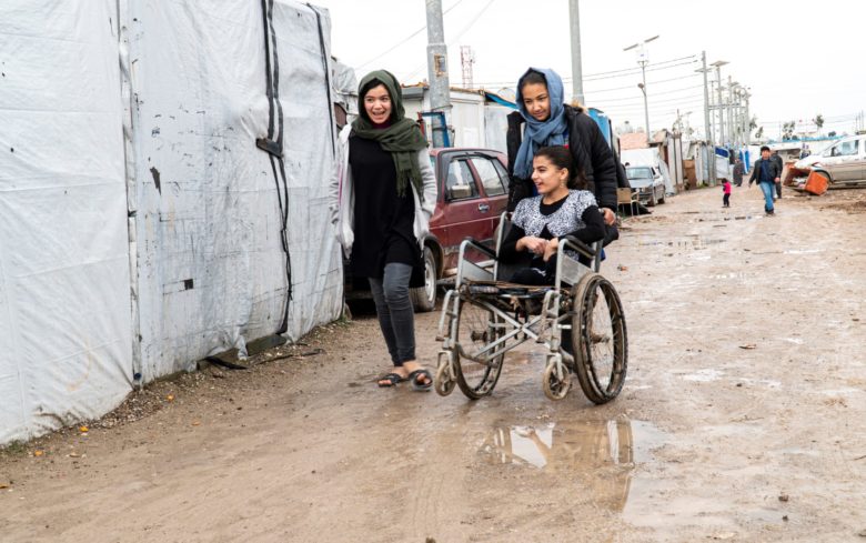 Kolme tyttöä mutaisella kadulla. Kaksi tytöistä kävelee, yksi tytöistä on pyörätuolissa, jota toinen tytöistä työntää. Pyörätuolissa olevalla tytöllä ei ole jalkoja.