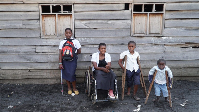 Neljä lasta harmaan rakennuksen edessä. Yksi lapsi istuu pyörätuolissa. Kahdella lapsista on kainalosauvat. Neljännellä lapsella on kävelykeppi.
