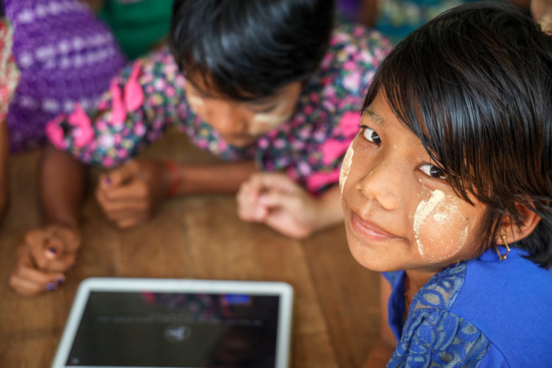 Myanmarilaisia lapsia pöydän ääressä kerääntyneenä tabletin ympärille.