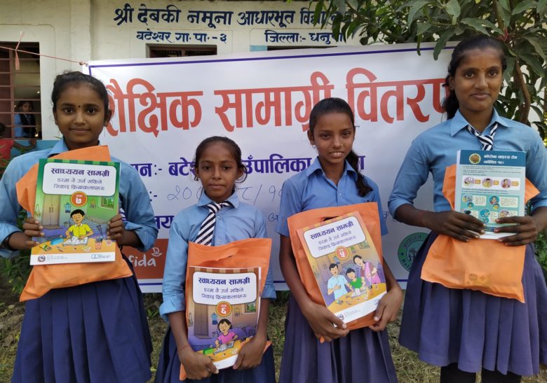 Neljä nepalilaista tyttöä seisoo koulupuvuissa koulun edessä. Tytöillä on kaikilla kädessään oppikirjat ja oranssi pussi.