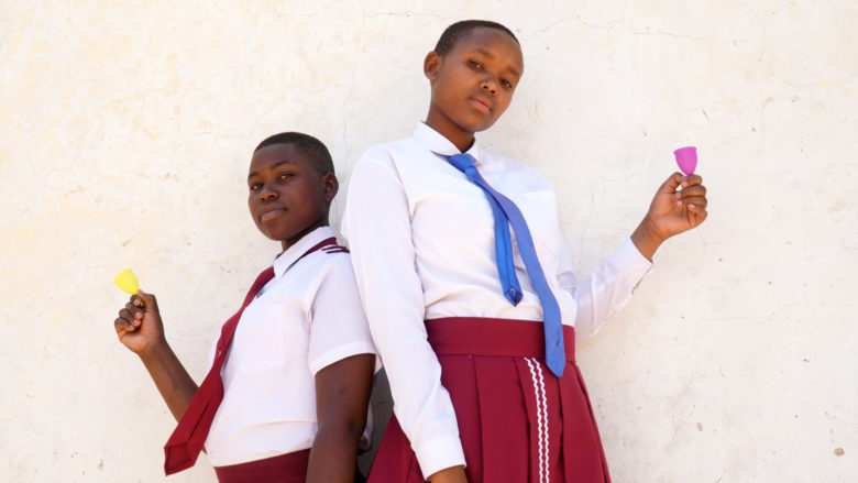Kaksi tyttöä seisoo seinän edessä koulupuvuissa. Molemmilla tytöillä on kädessään kuukuppi.