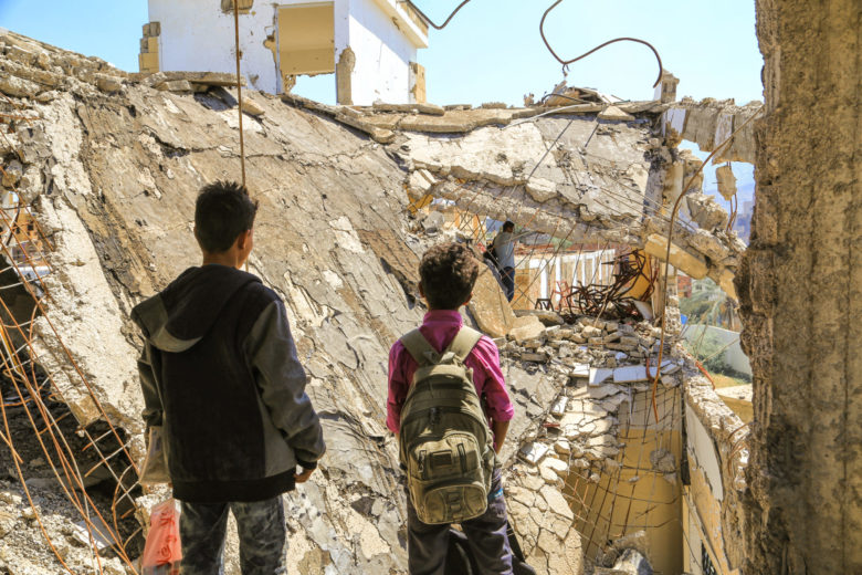 kaksi poikaa seisoo selin kameraan ja katsoo talon raunioita Jemenissä.
