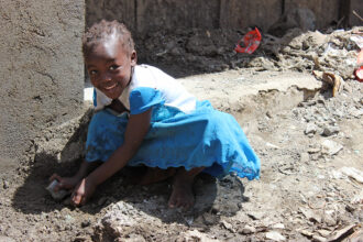 Pieni tyttö sinisessä mekossa leikkii maassa ja hymyilee.
