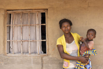 Kongolainen äiti keltaisessa paidassa seisoo seinän edessä lapsi sylissään.