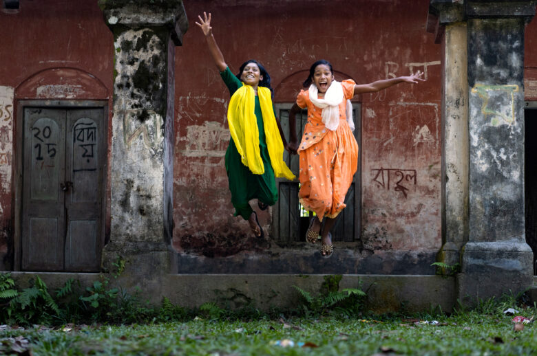 kaksi tummahiuksista tyttöä tunikaan pukeutuneena hyppäävät ilmaan. Toisella on keltainen huivi kaulassa ja toisella oranssi mekko.