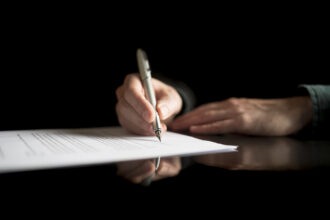 Lähikuva kädestä pöydällä, joka allekirjoittaa asiakirjaa.