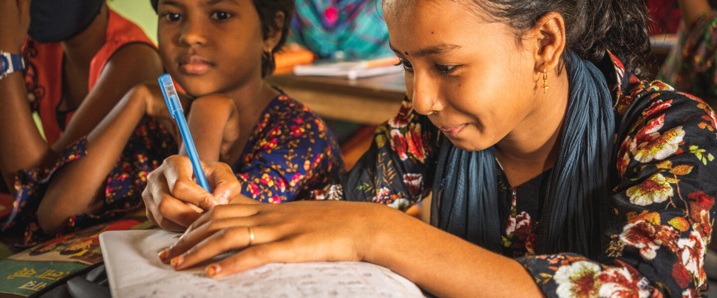 Bangladeshilainen tyttö kirjoittaa koulussa vihkoon ja hymyilee.