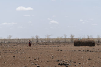 Kuvassa erämaata Kenian Marsabitin alueella, missä on pitkään kärsitty ilmastonmuutoksen aiheuttamasta kuivuudesta. Nainen kävelee kuvassa kohti maanviljelyä varten rakennettua varjorakennelmaa.